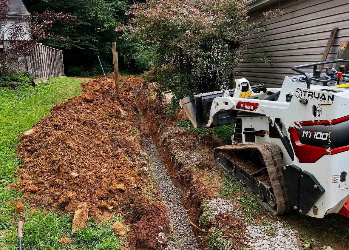 Mini track loader beside gravel-lined trench during drainage installation